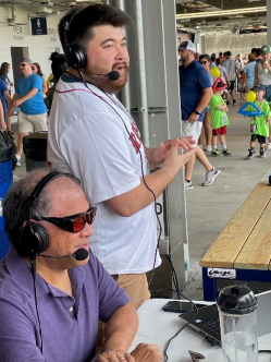 Blair Wong (seated) and son Evan Wong, both wearing headphone microphones, on the concourse, calling a WooSox game in 2022. Blair is the 2025 International Optician of the Year.